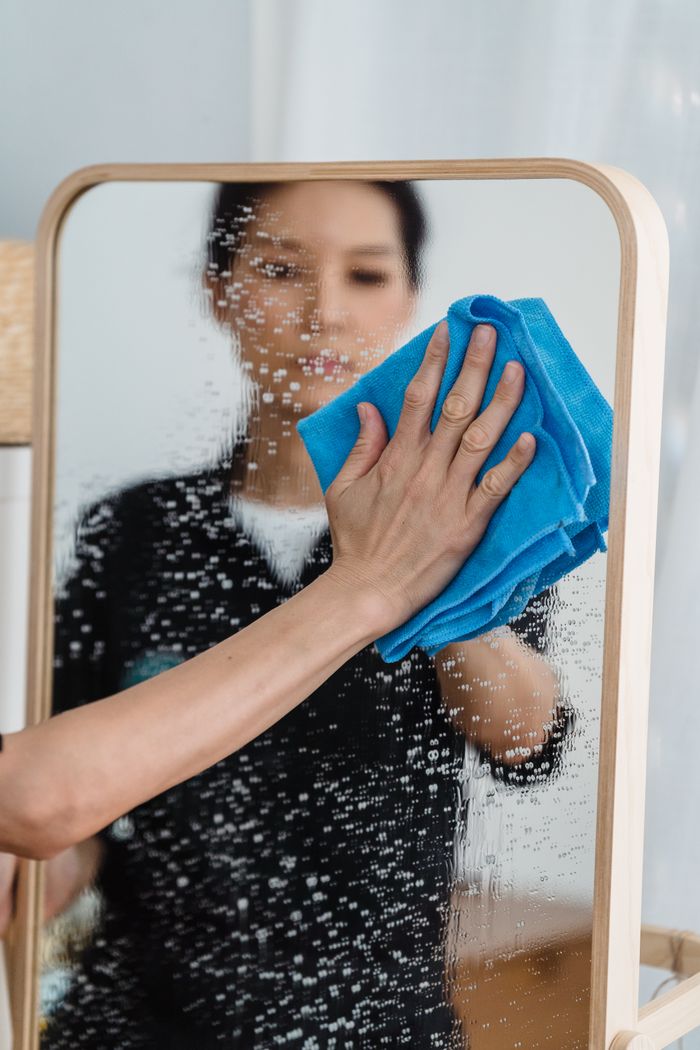 about-us-01 Close-up of a person wiping a mirror with a blue cloth, showing hands and reflection.