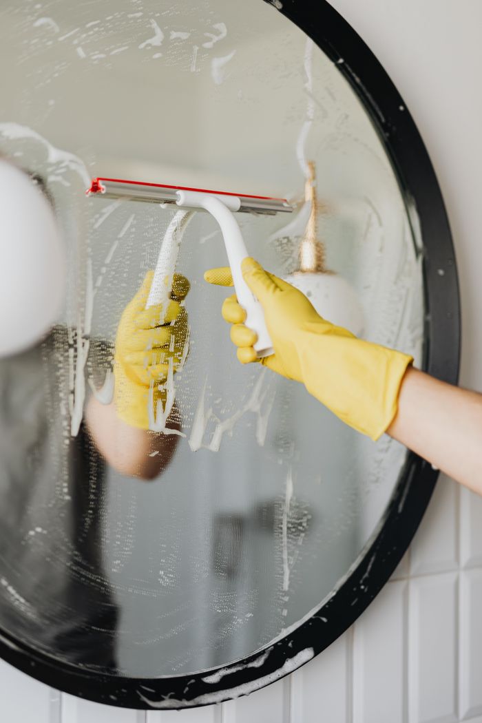 about-us-02 Faceless housekeeper in yellow rubber gloves cleaning round mirror from foam by squeegee in bathroom